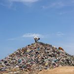 A man stands atop a mound of garbage under a bright blue sky, highlighting pollution.