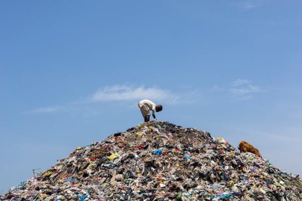 A man stands atop a mound of garbage under a bright blue sky, highlighting pollution.