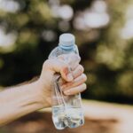 Crop unrecognizable man crumpling plastic bottle of water in hand against blurred background of green park trees on sunny summer day