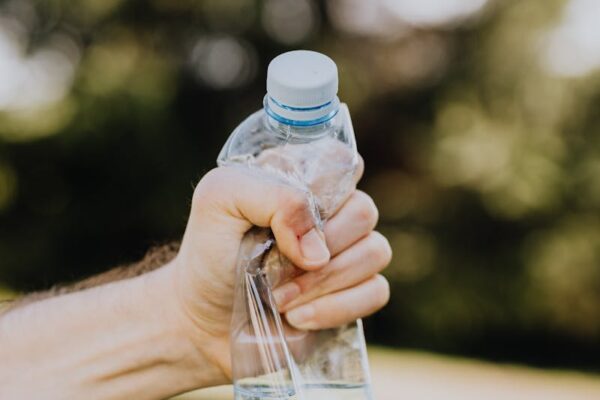 Crop unrecognizable man crumpling plastic bottle of water in hand against blurred background of green park trees on sunny summer day