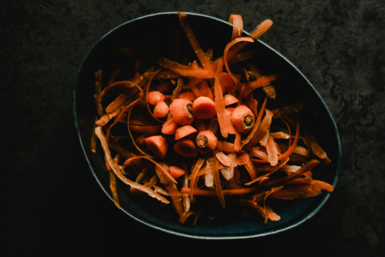 Close-up photo of fresh carrot peels and scraps in a dark bowl, perfect for food waste or preparation themes.