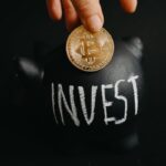 Gold bitcoin inserted into a black piggy bank labeled 'Invest', symbolizing cryptocurrency saving.