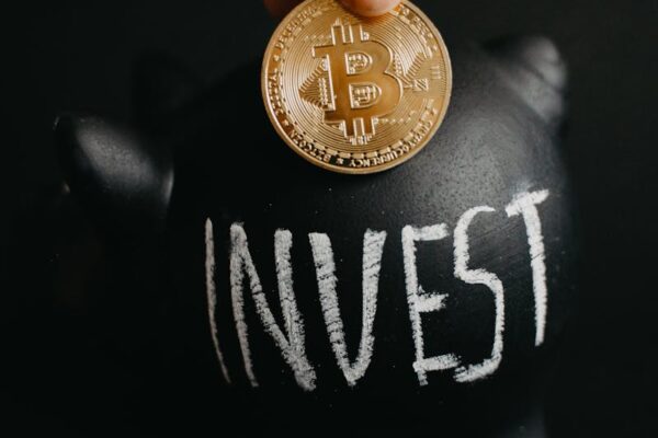 Gold bitcoin inserted into a black piggy bank labeled 'Invest', symbolizing cryptocurrency saving.
