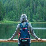 A person in a hoodie stands by a serene lake surrounded by lush greenery, enjoying the peaceful outdoors.