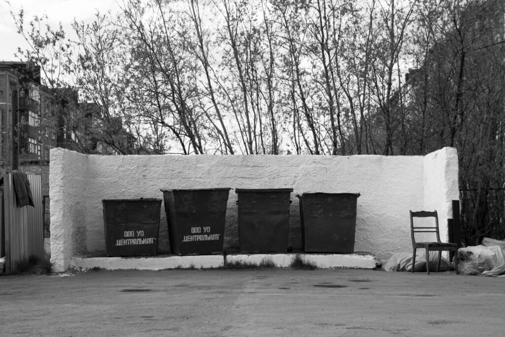 Monochrome photo of urban garbage bins and a solitary chair in a residential area.