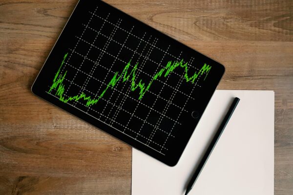 Flatlay of an iPad displaying stock market graph on a wooden desk with a pencil and paper.