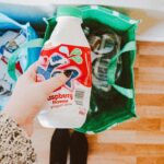 A person recycling a raspberry yogurt drink bottle into a green recycling bag.