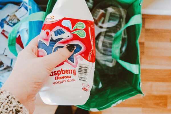A person recycling a raspberry yogurt drink bottle into a green recycling bag.