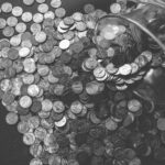 Monochrome image of coins spilling from a glass jar, symbolizing wealth and savings.