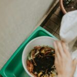 View of hands adding food scraps to a compost bin with visible soil.