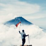 Hiker atop Sindoro Mountain in Central Java, Indonesia, with flag under vibrant sky.