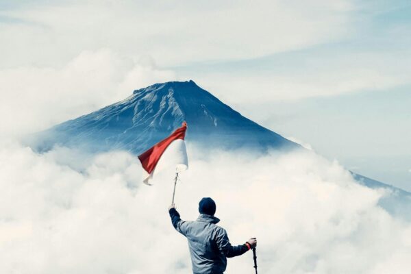Hiker atop Sindoro Mountain in Central Java, Indonesia, with flag under vibrant sky.