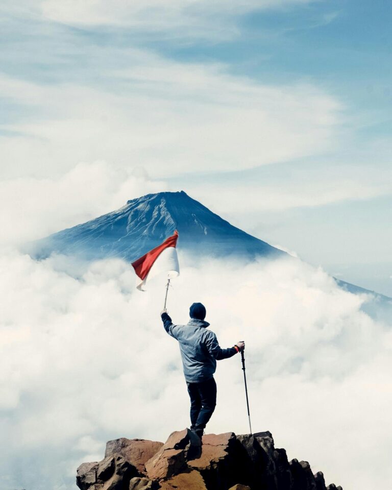 Hiker atop Sindoro Mountain in Central Java, Indonesia, with flag under vibrant sky.