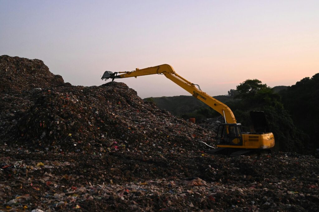 Yellow excavator operates amidst piles of waste in a Bangladesh landfill at sunset.