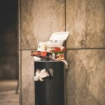 A trash can overflowing with garbage in an urban setting, against a concrete wall.