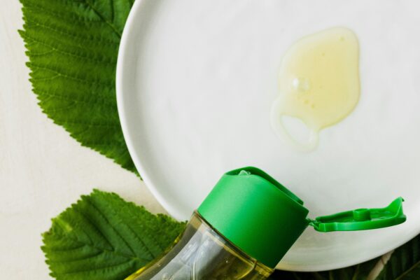 Top view of open bottle of cosmetic oil placed flat on table near white plate with small amount of oil on it on big fresh green leaf