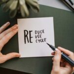 Close-up of hands writing a motivational 'Reduce Reuse Recycle' note on paper with a pen, surrounded by office items.