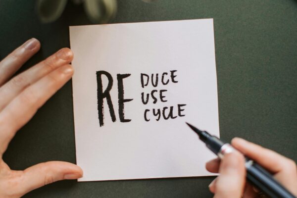 Close-up of hands writing a motivational 'Reduce Reuse Recycle' note on paper with a pen, surrounded by office items.