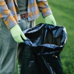 Person wearing gloves holding a trash bag in a green field, emphasizing volunteer cleanup efforts.