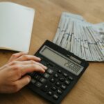 A hand calculates financial figures using a calculator with stacks of cash nearby on a wooden table.