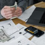 Hands counting money at a workspace with a laptop, calculator, and financial documents.
