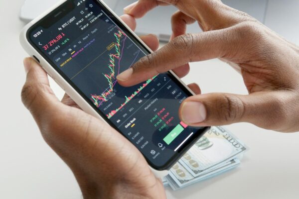 Hands using smartphone trading app with charts, dollar bills in background.