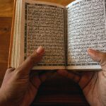 A close-up of hands holding an open Quran on a wooden table, showcasing Arabic script.