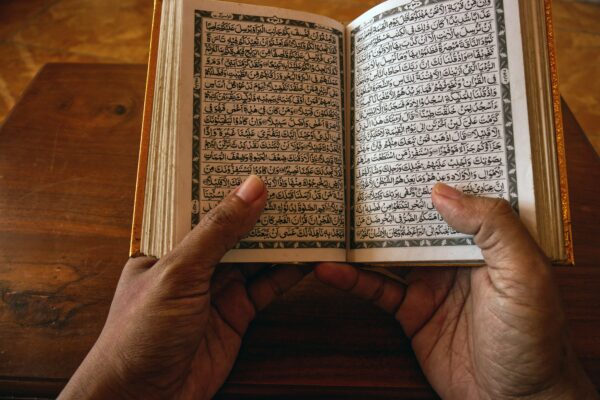 A close-up of hands holding an open Quran on a wooden table, showcasing Arabic script.