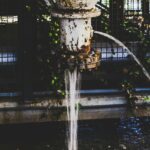 A rusty pipe discharges water into an industrial environment, surrounded by lush foliage.