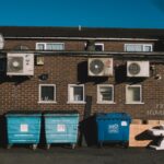 Exterior view of urban alley with brick wall, recycling bins, and air conditioning units.