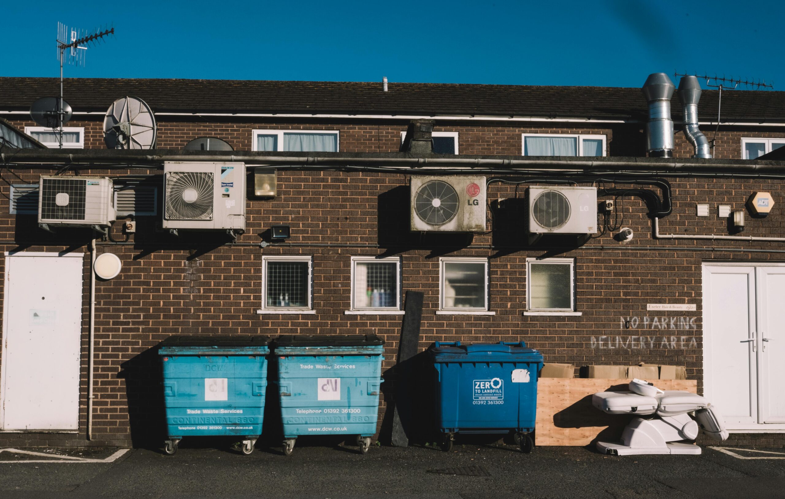 Exterior view of urban alley with brick wall, recycling bins, and air conditioning units.