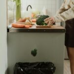Person discarding broccoli in a stylish kitchen with fresh vegetables on the counter.