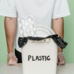 Adult male with gloves standing by a plastic recycling bin full of waste.