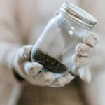 Hands in gloves holding a glass jar containing soil, emphasizing texture and detail in a close-up, blurred background.