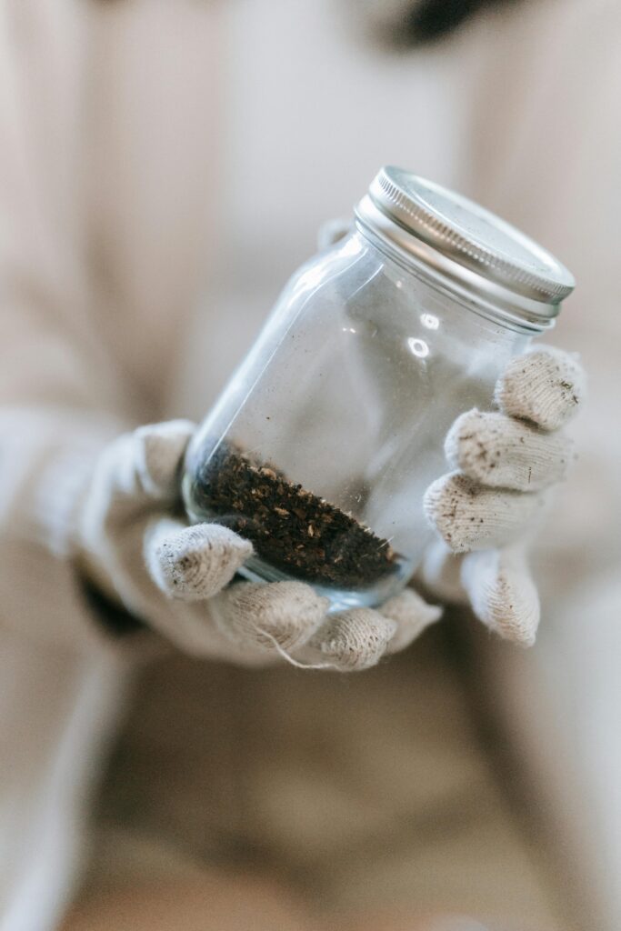 Hands in gloves holding a glass jar containing soil, emphasizing texture and detail in a close-up, blurred background.