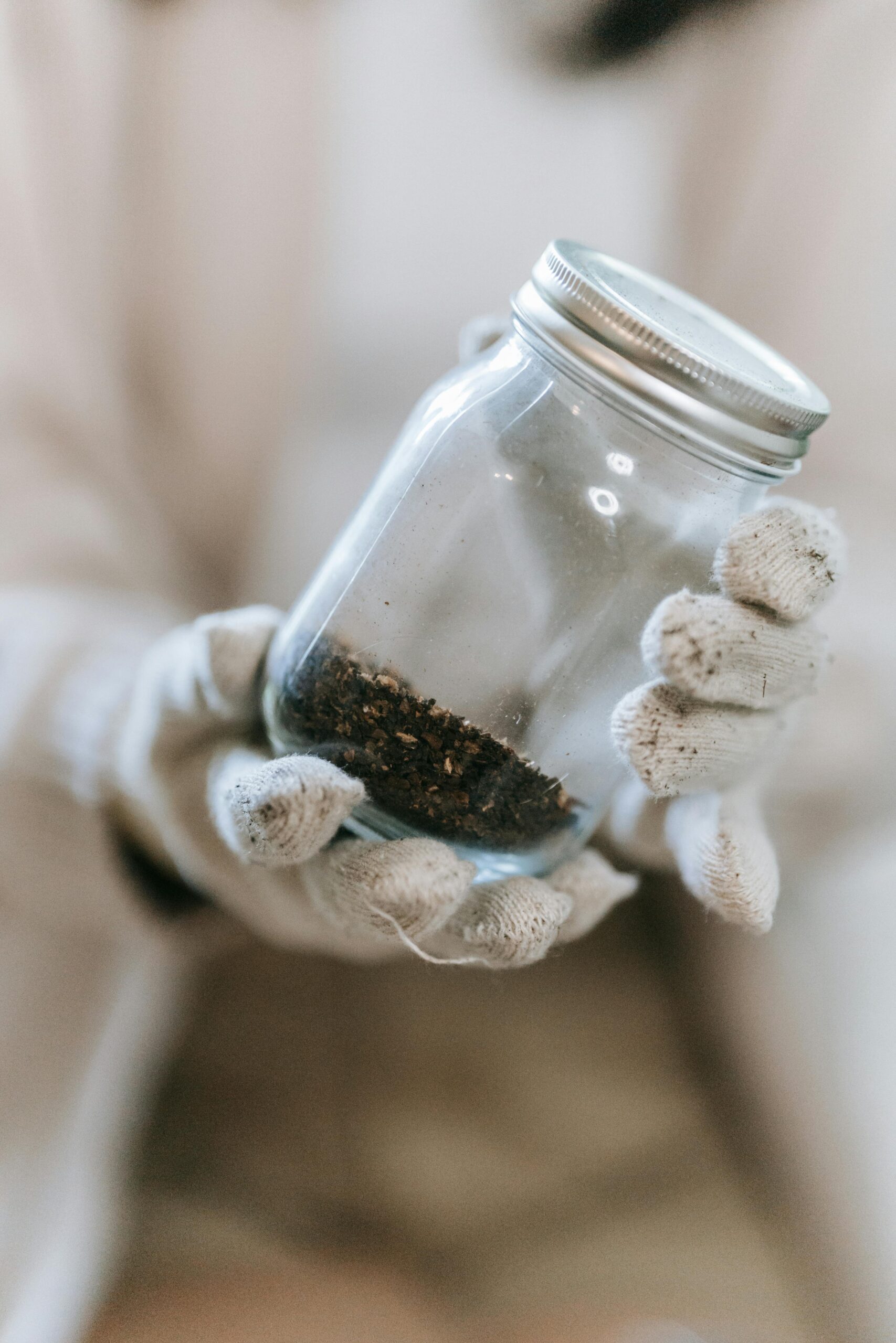 Hands in gloves holding a glass jar containing soil, emphasizing texture and detail in a close-up, blurred background.
